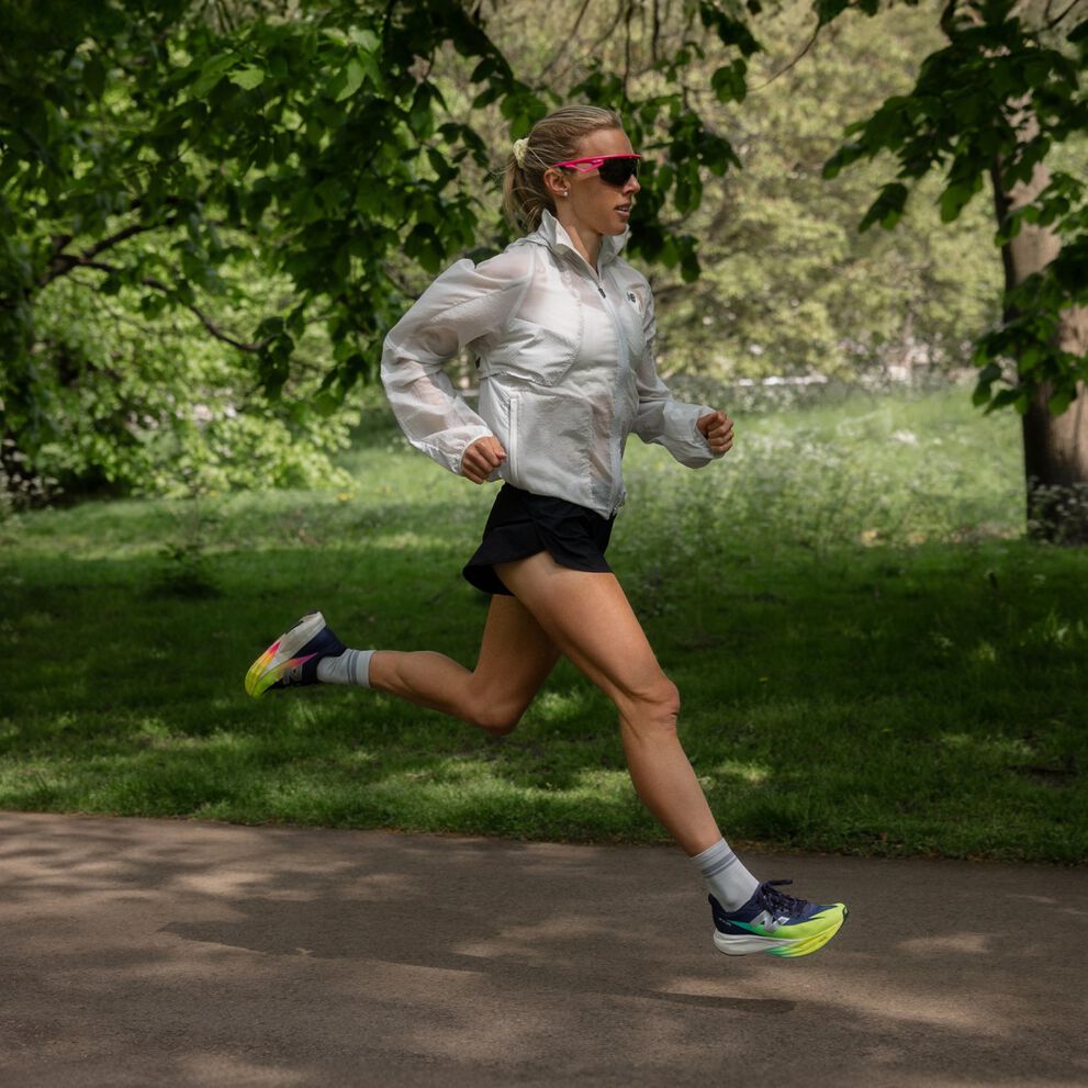 Emily Sisson running outdoors on a paved path in a park, wearing a white jacket, black shorts, and brightly colored running shoes. 
