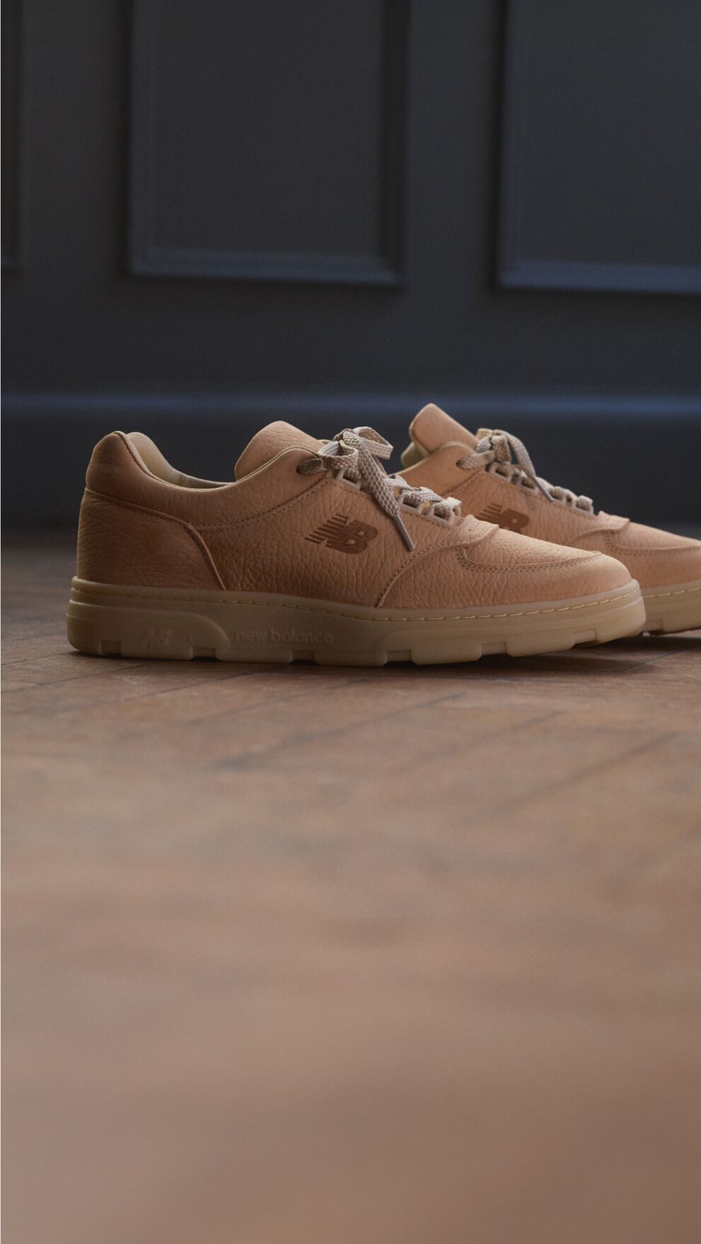 A pair of New Balance Made in UK sneakers on a wooden floor with dark paneled walls in the background.