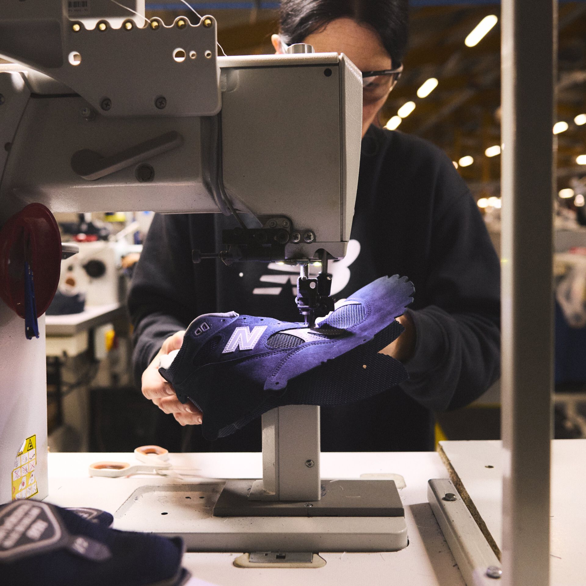 A worker in the New Balance Made in UK factory carefully sewing together a sneaker upper in an industrial environment with a sewing machine.