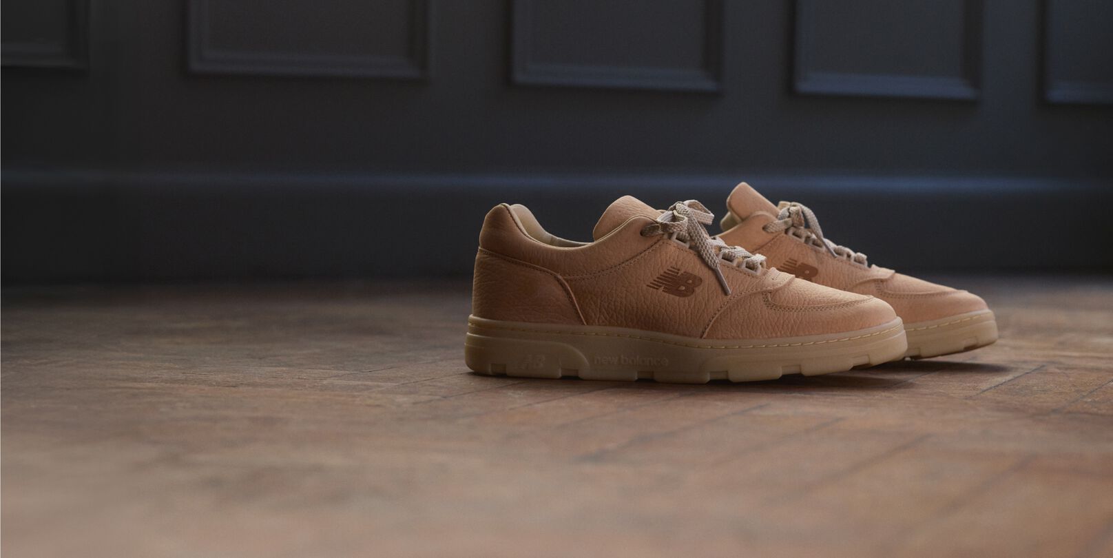 A pair of New Balance Made in UK sneakers on a wooden floor with dark paneled walls in the background.